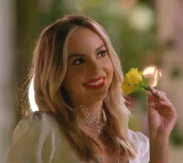 A woman with long blonde hair and a bright smile holds a yellow rose, set against an evening background.