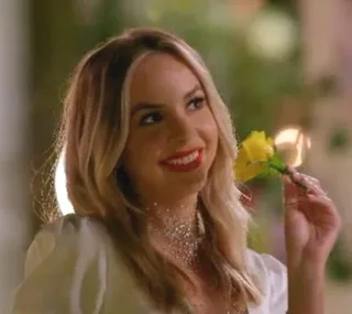 A woman with long blonde hair and a bright smile holds a yellow rose, set against an evening background.