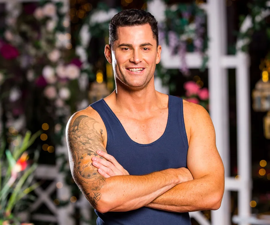 Man with dark hair, smiling, arms crossed, sleeveless shirt, tattoo on right arm, vibrant floral background.