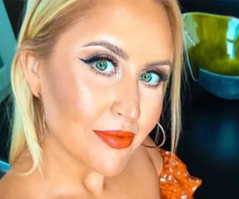 Blonde woman with green eyes and orange lipstick wears an orange top, posing indoors with a confident expression.