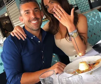 A couple celebrating at a restaurant; the woman shows an engagement ring, smiling with a dessert and sparkler on the table.