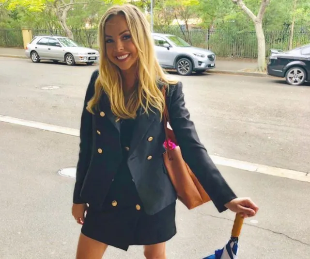 A woman in a black blazer and skirt smiles while holding an umbrella and a brown bag, standing on a street.
