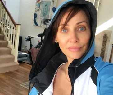 A person in a blue and black hooded jacket taking a selfie in a home with exercise equipment in the background.