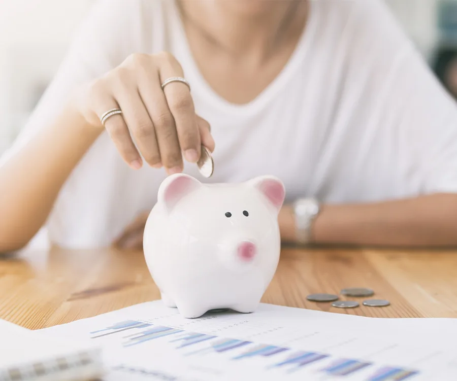 Person putting coin into a white piggy bank with charts and coins on the table.