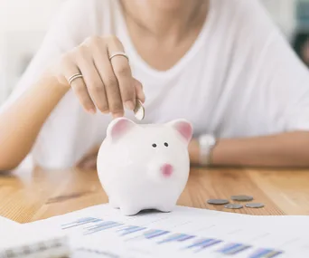 Person putting coin into a white piggy bank with charts and coins on the table.
