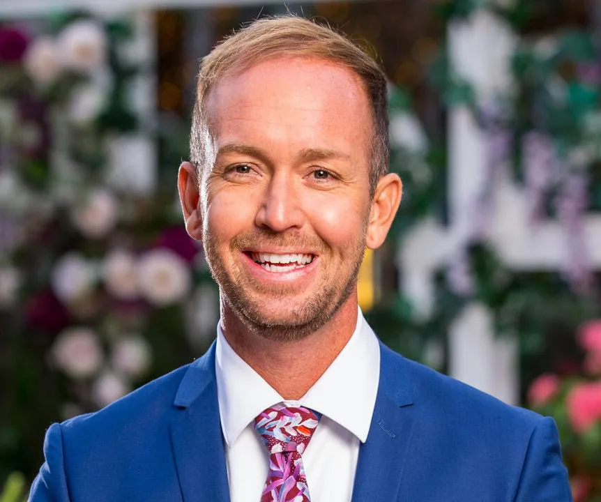 Man in a blue suit smiling, wearing a colorful tie, with a floral backdrop.