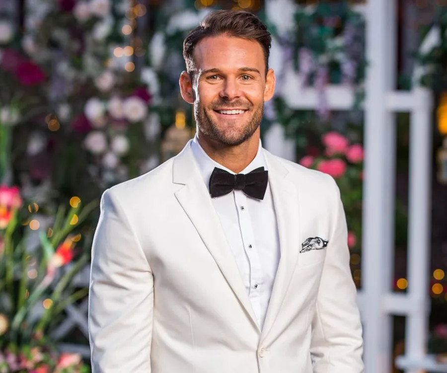 A man in a white tuxedo with a black bow tie smiles, standing in front of a floral background.