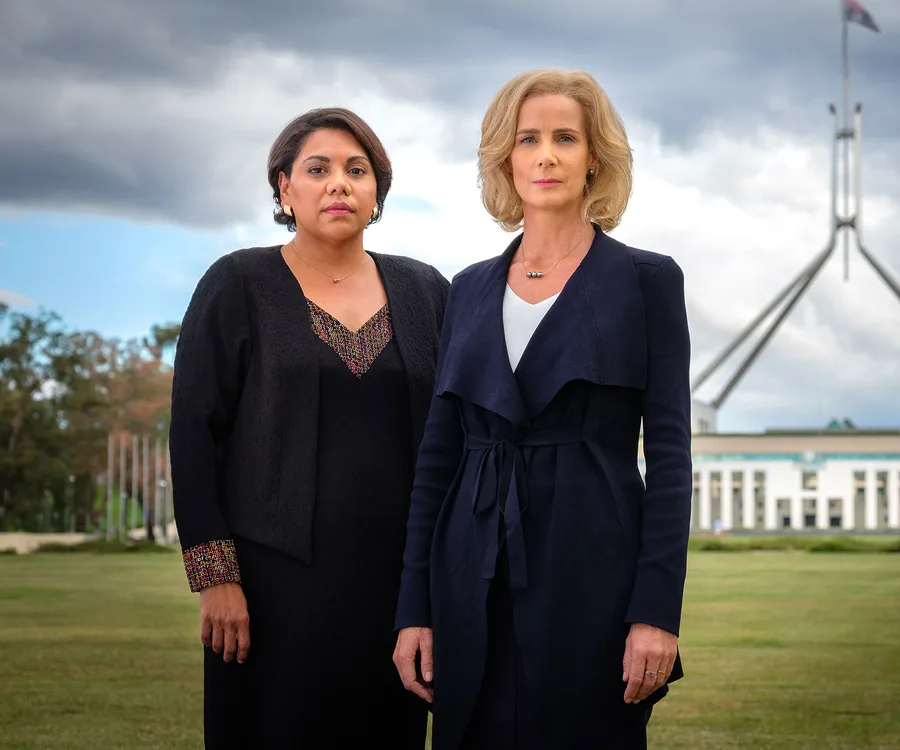 Two women stand on a lawn in front of a large building under a cloudy sky, wearing professional attire and looking serious.