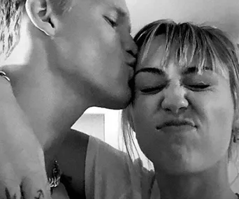 A black and white photo of a person kissing another on the side of their face, both looking playful and affectionate.