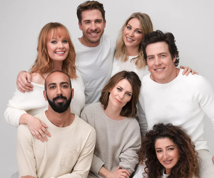 Seven people in light-colored casual wear are smiling, some embracing each other, posing against a plain background.