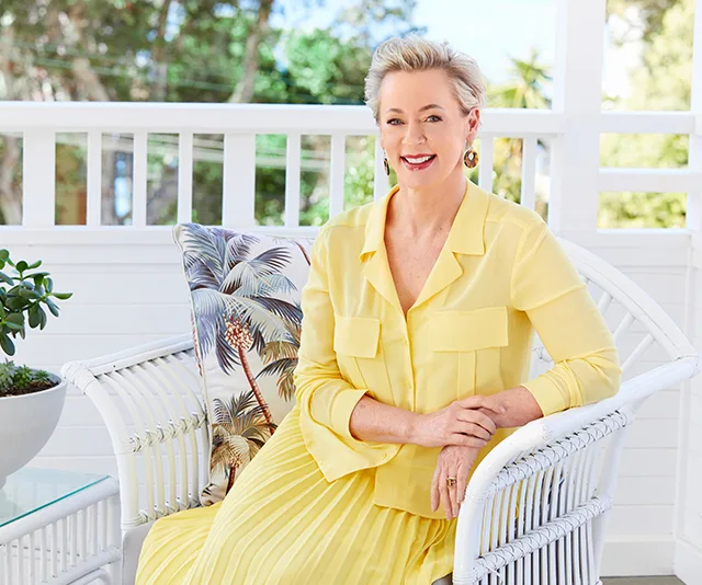 Woman in a yellow outfit sitting on a white wicker chair with a tropical pillow, smiling on a sunny porch.