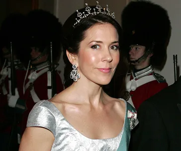 Princess Mary wearing a tiara and earrings, standing in front of royal guards in uniform.