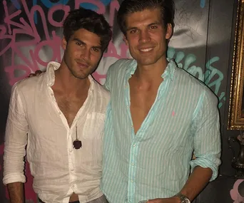 Two men smiling in front of graffiti wall, one in a white shirt, the other in a light striped shirt.
