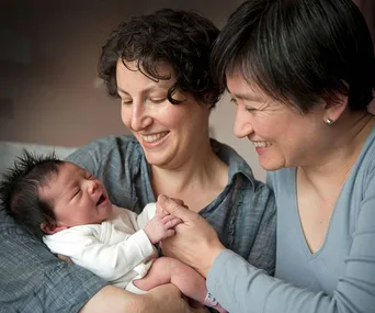Two women smiling at each other while holding a newborn baby in their arms.
