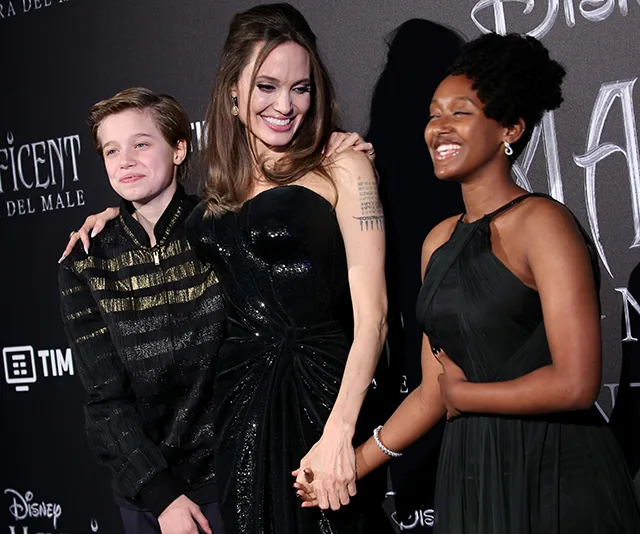 Three people smiling at a formal event; one in a black dress holding hands, standing against a "Maleficent" backdrop.