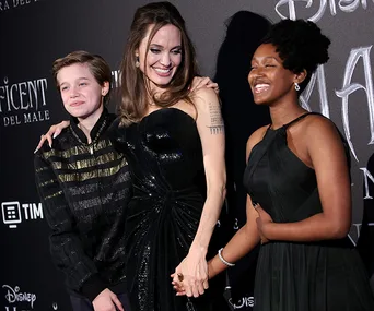 Three people smiling at a formal event; one in a black dress holding hands, standing against a "Maleficent" backdrop.