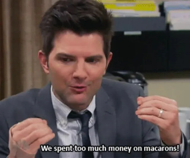 A man in a suit gestures with hands, saying, "We spent too much money on macarons!" in a humorous scene.