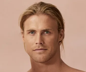 A person with long blond hair and a neutral expression against a soft pink background.