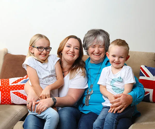 Four people, including an elderly woman and two children, sit smiling on a couch with decorative pillows.