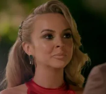 A woman with long blonde hair and earrings in a red outfit, appearing focused, likely during a dramatic moment.