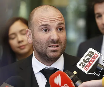 A man in a suit speaks into multiple microphones with people in the background.