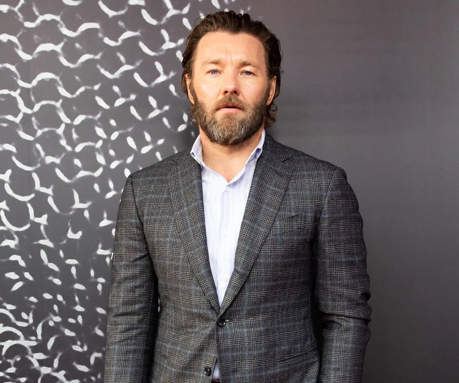A man in a checkered suit and striped shirt stands against a patterned background.