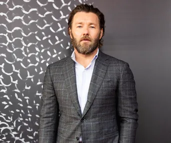 A man in a checkered suit and striped shirt stands against a patterned background.