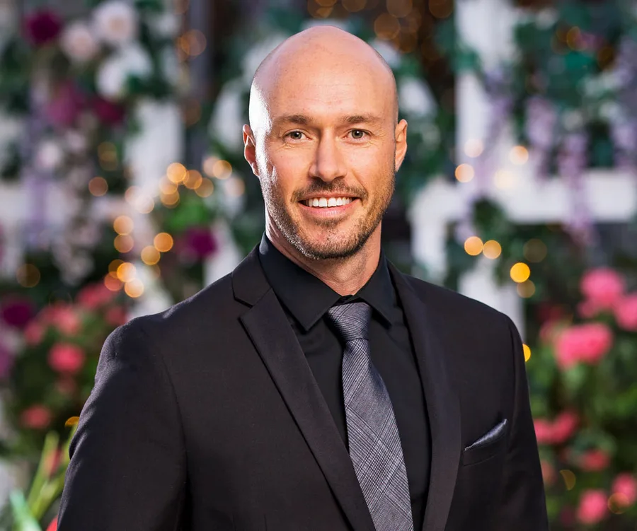 Man in a suit, smiling, with a blurred floral and twinkle light background.