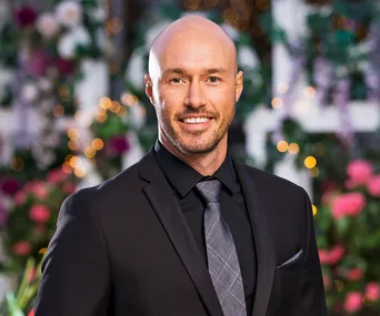 Man in a suit, smiling, with a blurred floral and twinkle light background.