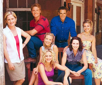 Cast members from "McLeod's Daughters" pose together outside a rustic building. They are smiling and relaxed.