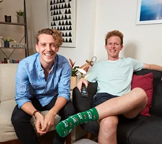 Two smiling men on a sofa, one in a blue shirt, the other in a striped shirt with festive socks, holding a remote.