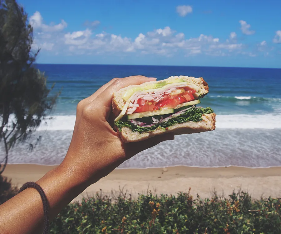 Hand holding a sandwich with veggies and turkey against a beach and ocean backdrop under a bright blue sky.