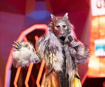 A performer in a detailed wolf costume sings on stage with vibrant lighting in the background.