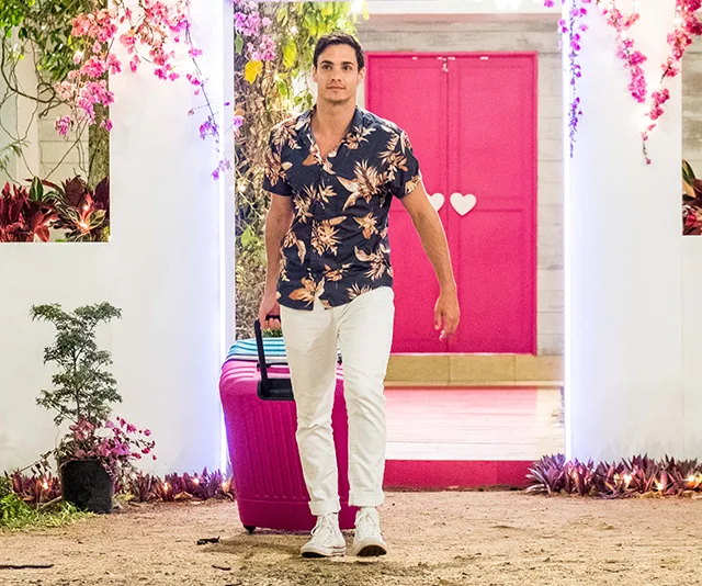 Man in floral shirt and white pants walks with pink suitcase near pink door, surrounded by flowers.