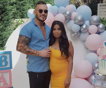 A couple stands in front of pastel balloons at a baby shower; the man wears sunglasses and the woman is in a yellow dress.