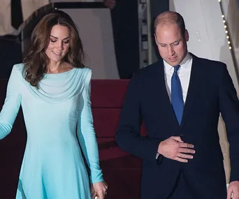 A man in a suit and a woman in a light blue dress descend red-carpeted stairs, possibly from an aircraft.