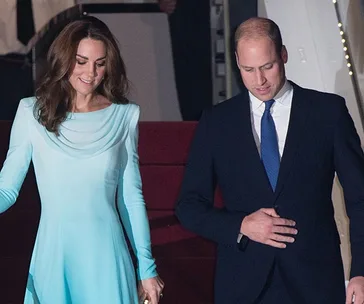 A man in a suit and a woman in a light blue dress descend red-carpeted stairs, possibly from an aircraft.