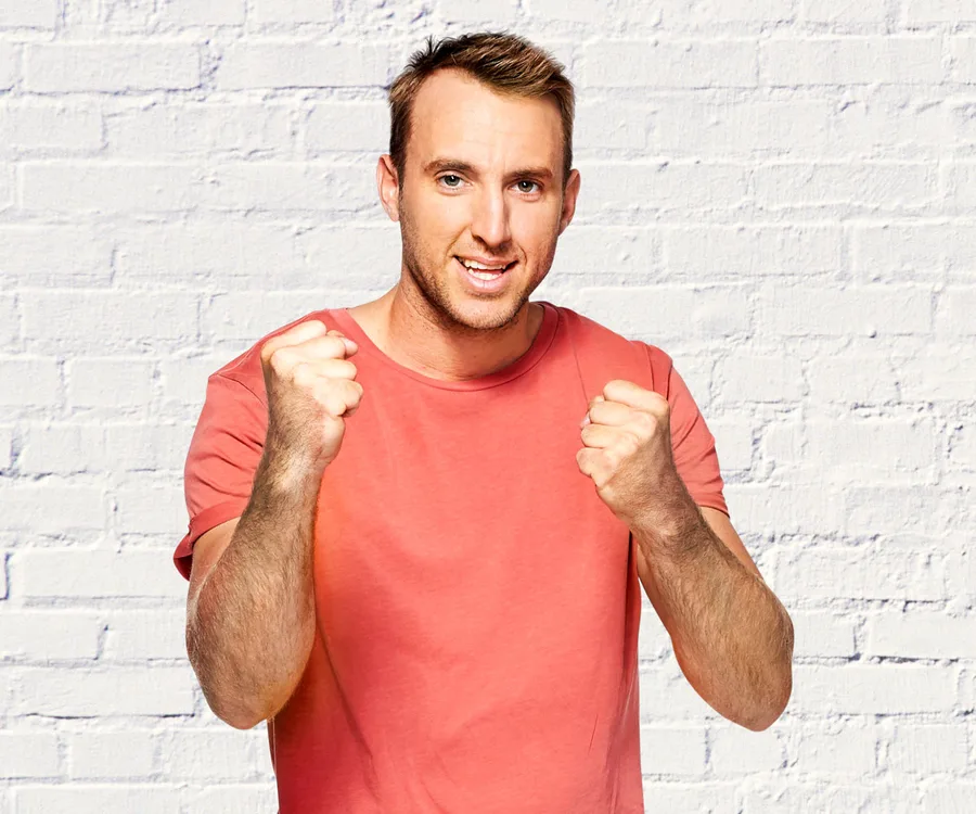Man in a red shirt with fists raised, posing against a white brick wall background.