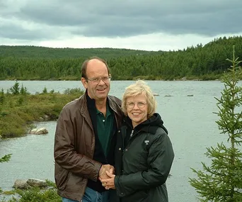 Couple standing by a serene lake with lush forests in the background, smiling under a cloudy sky.