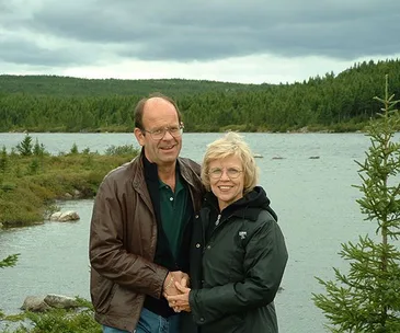Couple standing by a serene lake with lush forests in the background, smiling under a cloudy sky.