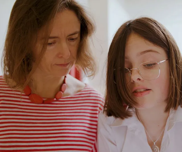 A woman in a red-striped shirt and a girl with glasses in a white top look at something together, focused and engaged.
