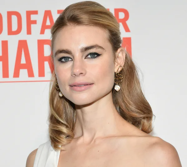 Lucy Fry at a "Godfather of Harlem" event, wearing elegant earrings and a white dress.
