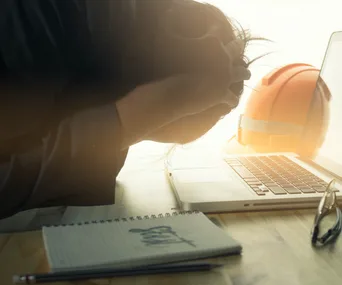 Man holding head in hands, overwhelmed at desk with laptop, notebook, and orange hard hat.