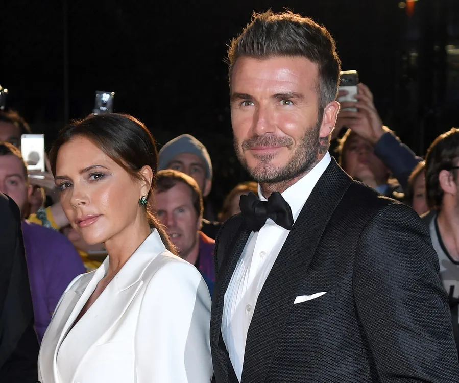 Victoria and David Beckham in formal attire at a public event, with people in the background taking pictures.
