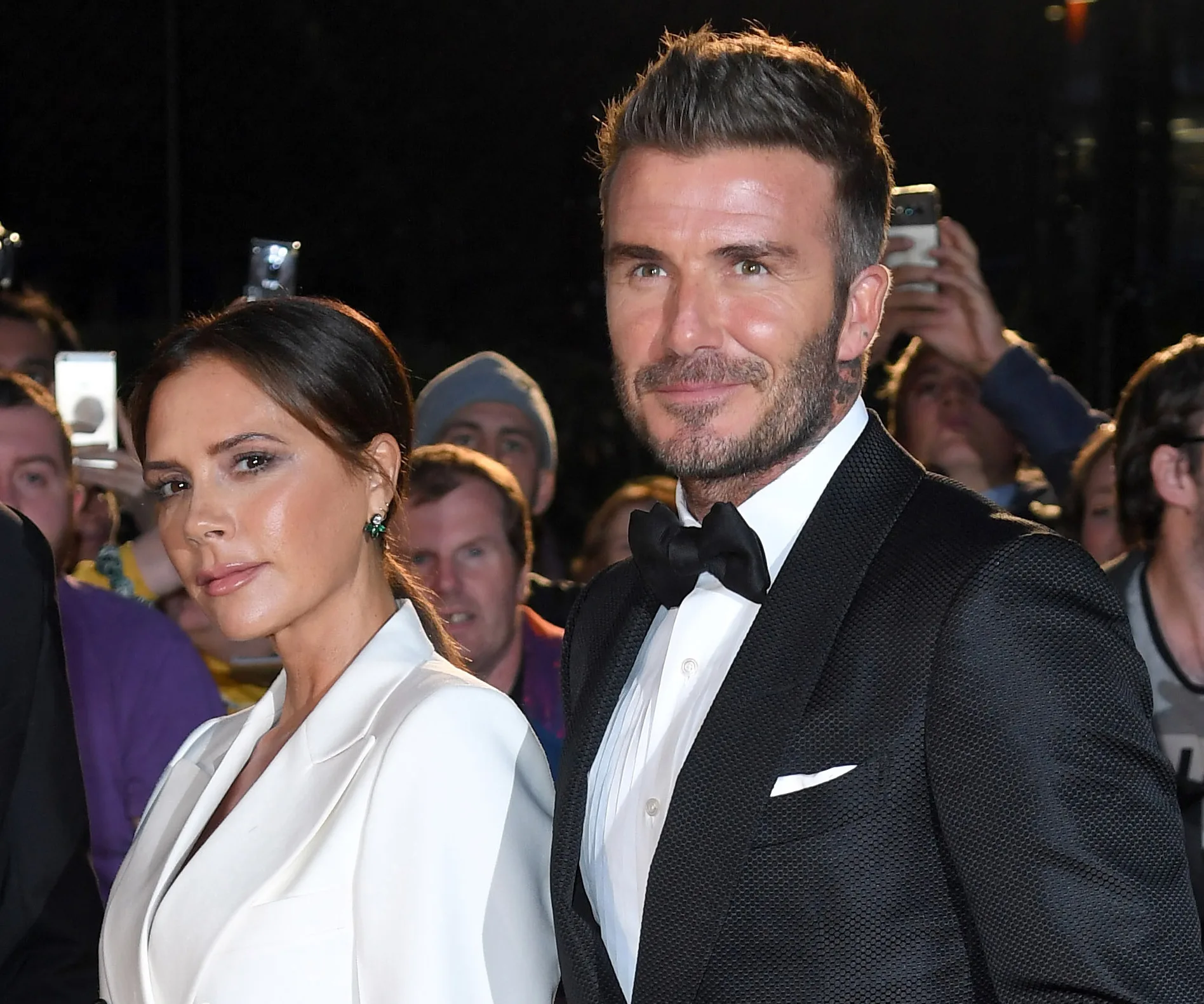 Victoria and David Beckham in formal attire at a public event, with people in the background taking pictures.