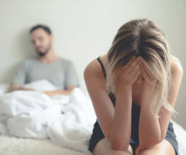 Woman sitting in distress on bed, man behind her looking away, emotional tension evident.