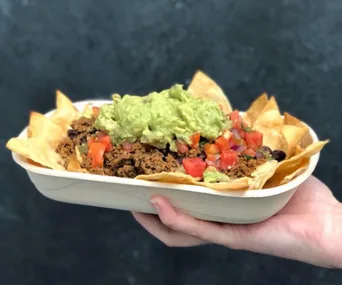 A hand holding a tray of nachos with guacamole, tomatoes, and ground meat, set against a dark background.