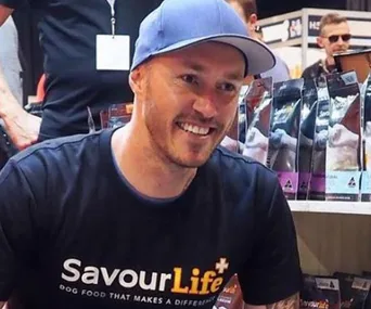 Man in a blue cap and SavourLife shirt smiling, standing near pet food products.