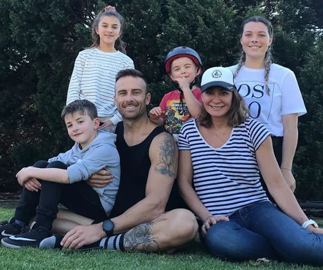 A smiling family sits on grass in front of trees, with two adults and four children, one wearing a helmet.