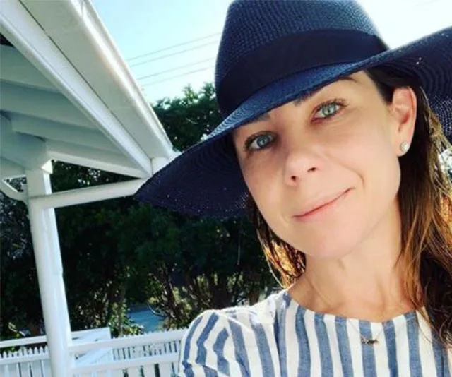A woman in a striped shirt and wide-brimmed hat takes a selfie outdoors near white railing and trees.